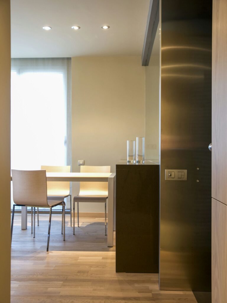 Área de Comedor Minimalista Moderna con Luz Natural Un área de comedor elegante y minimalista que presenta una mesa cuadrada blanca, sillas de tonos claros y suelo de madera, iluminada por una suave luz natural que entra a través de cortinas ligeras.