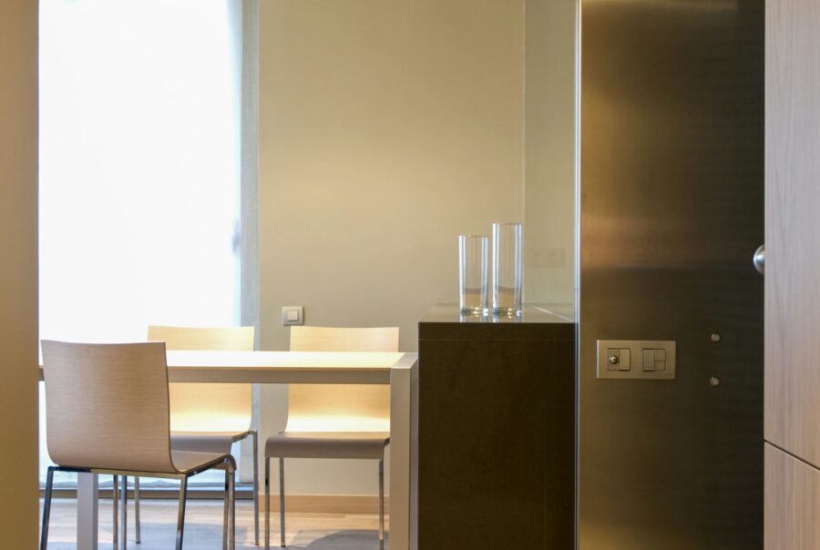Un área de comedor elegante y minimalista que presenta una mesa cuadrada blanca, sillas de tonos claros y suelo de madera, iluminada por una suave luz natural que entra a través de cortinas ligeras.