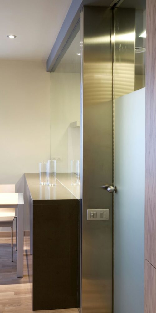 Una cocina contemporánea con suelos de madera clara, encimeras brillantes y una puerta de vidrio y metal. El espacio se complementa con una mesa minimalista rodeada de sillas blancas, con paredes beige y gabinetes oscuros, mostrando una estética limpia.