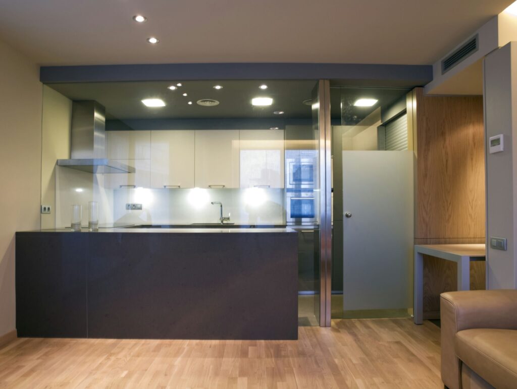 Cocina Moderna con Diseño Elegante y Electrodomésticos Contemporáneos Una cocina moderna y elegante con encimeras oscuras, armarios de colores claros y electrodomésticos de acero inoxidable, incluyendo una campana sobre la estufa. El espacio está iluminado por luces empotradas y tiene un suelo de madera que aporta calidez. A la derecha se puede ver una pequeña área de estar con una silla marrón claro, que contribuye a la estética minimalista de la cocina.