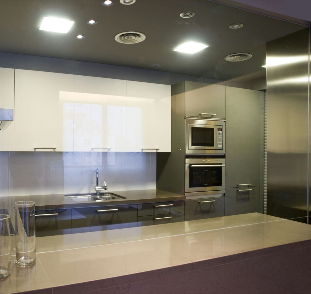 Diseño de cocina moderna y elegante con estética contemporánea Una cocina moderna con un diseño elegante que presenta armarios superiores blancos y un mobiliario inferior gris. Una encimera brillante se extiende junto al microondas y horno empotrados, y un fregadero ubicado en el centro. La iluminación empotrada brillante resalta las líneas limpias y el enfoque minimalista de la cocina.
