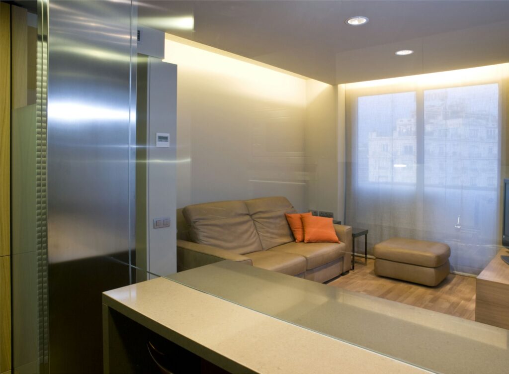 Espacio de Vida Moderno y Elegante con Diseño Minimalista y Vista Urbana Una sala de estar moderna con una encimera elegante con bordes curvados, un acogedor sofá de cuero marrón claro con un cojín naranja y un pequeño otomano. El espacio está bien iluminado con luz natural que se filtra a través de grandes ventanas adornadas con cortinas ligeras, mostrando una estética contemporánea.