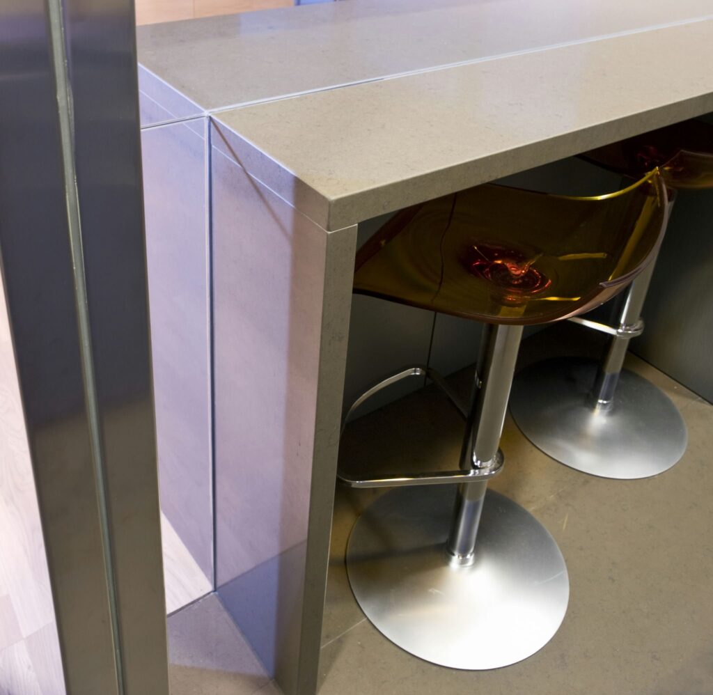 Cocina Moderna Minimalista con Encimeras Claras y Elegante Área de Bar Una cocina moderna que presenta una encimera clara y elegante con un acabado sin junta, junto a un área de bar contemporánea adornada con elegantes taburetes de bar ámbar transparentes con bases metálicas, mostrando un diseño minimalista y elegante.