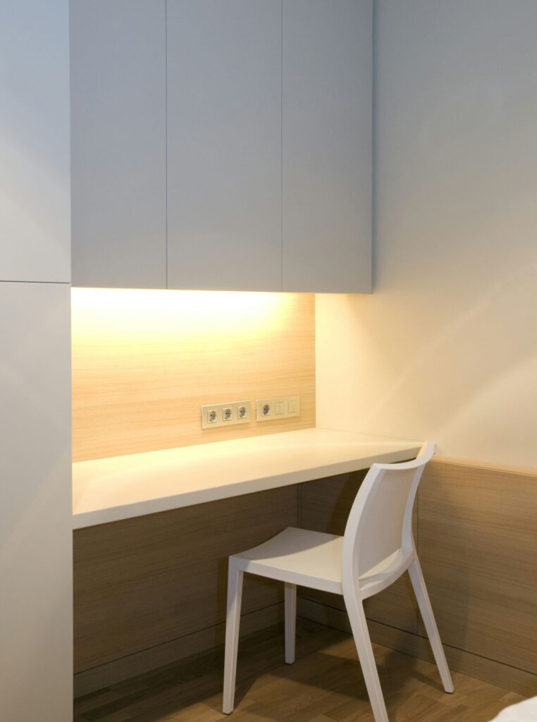 Espacio de trabajo moderno y minimalista con silla blanca y escritorio de color claro Un espacio de trabajo elegante y minimalista que cuenta con una silla blanca en un escritorio de color claro, iluminado por luz cálida de una lámpara de techo. El diseño incluye gabinetes de madera clara y enchufes visibles, creando una atmósfera serena perfecta para una oficina en casa o un área de estudio.