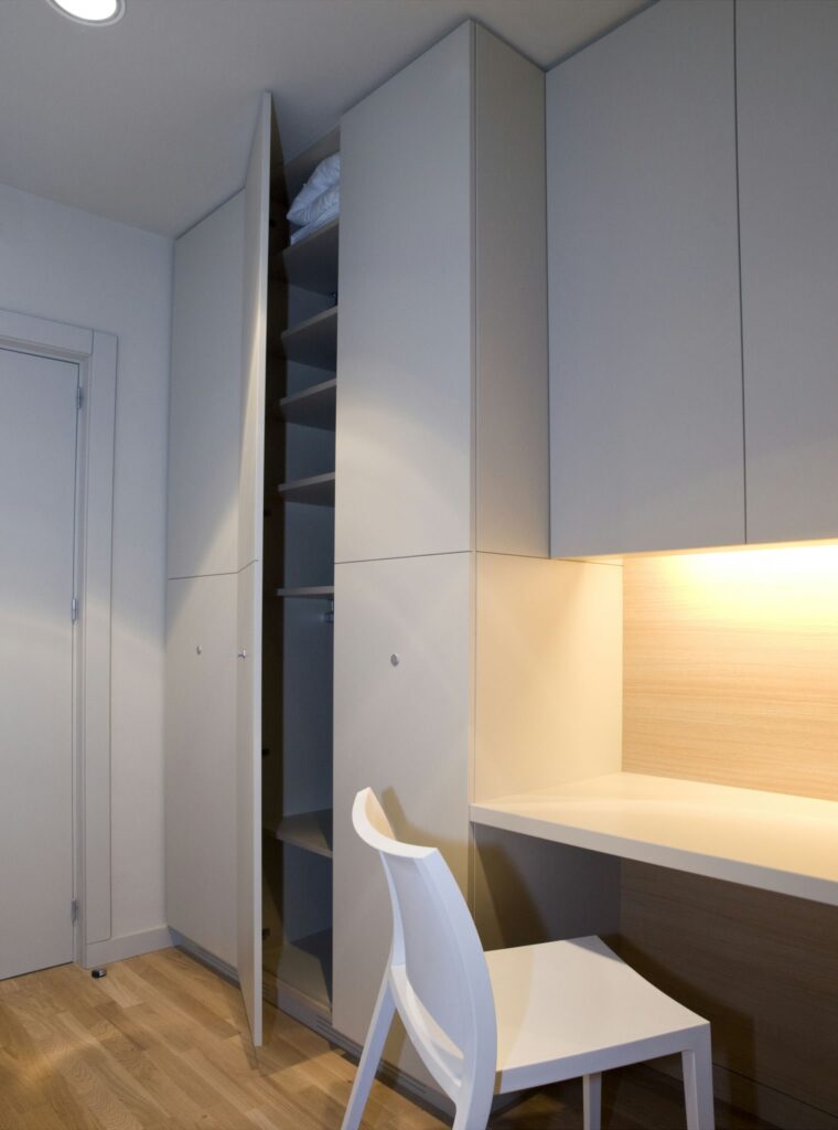 Habitación Modernista Minimalista con Mobiliario Empotrado y Espacio de Trabajo Una habitación modernista minimalista que cuenta con mobiliario empotrado, incluyendo un armario alto y estrecho con estantes abiertos y una mesa clara iluminada por cálidas luces de techo, creando una atmósfera acogedora con suelo de madera clara y paredes neutras.