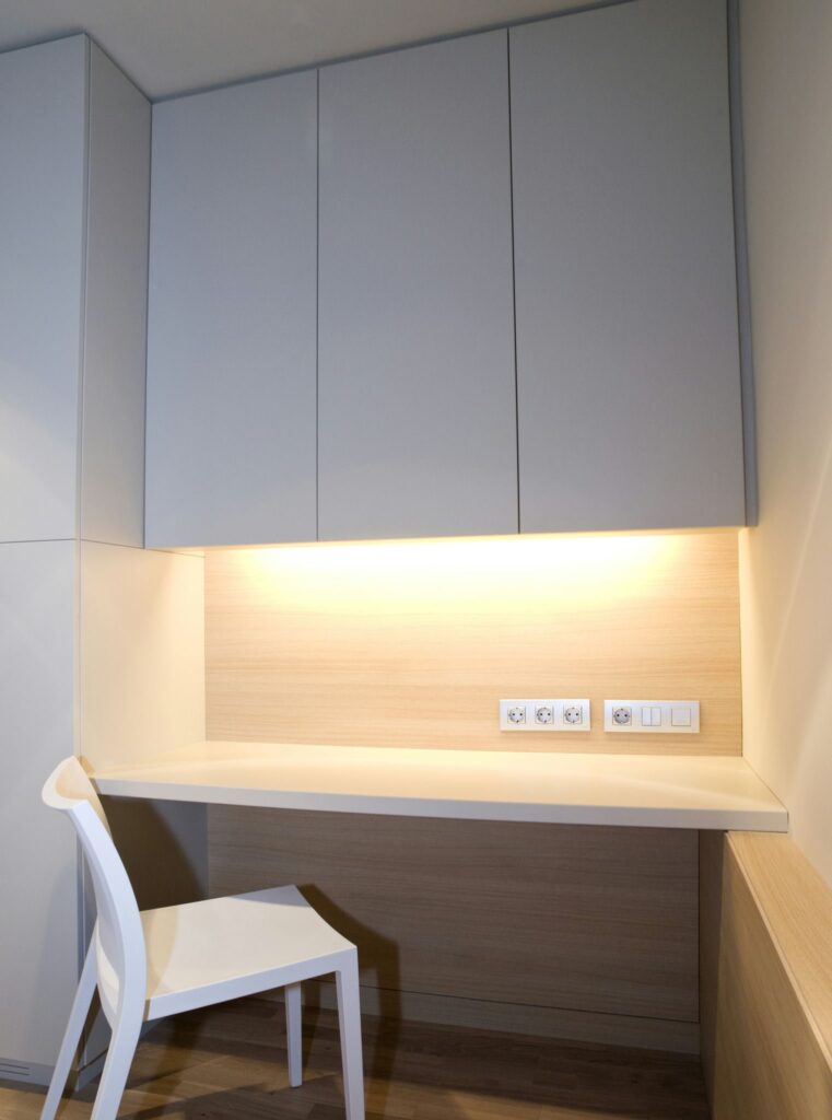 Espacio de Trabajo Moderno y Minimalista con Escritorio Montado en la Pared y Soluciones de Almacenamiento Elegantes Un espacio de trabajo elegante y minimalista que presenta un escritorio montado en la pared de color claro complementado por gabinetes grises elegantes y una silla blanca simple. La iluminación empotrada añade calidez al área, con tomacorrientes visibles para un fácil acceso a la energía.