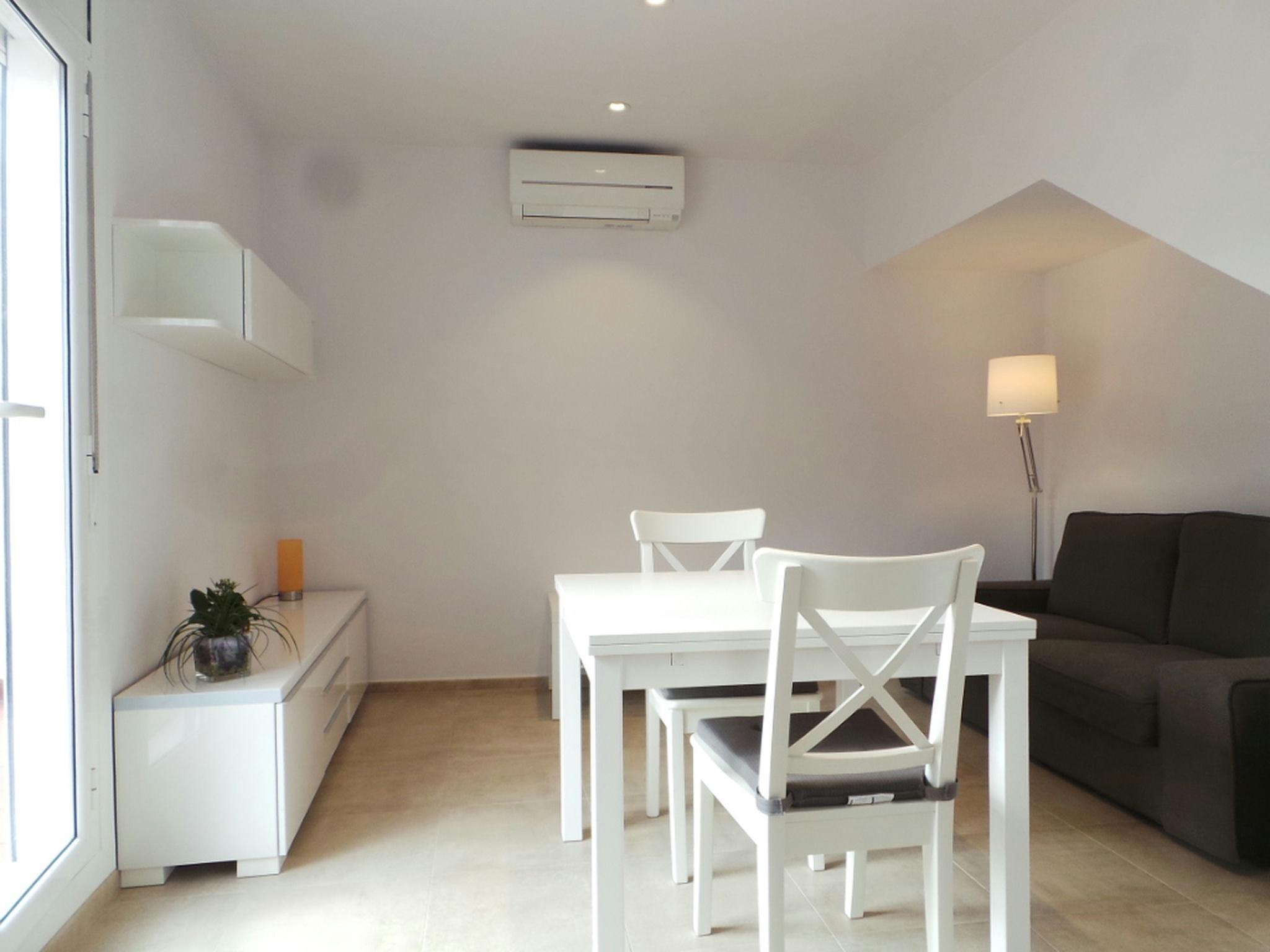 Interiores de un comedor contemporáneo en Barcelona, con una mesa blanca minimalista, sillas elegantes, un sofá gris oscuro y un mueble de almacenamiento blanco, iluminado por luz natural y decorado con una planta.