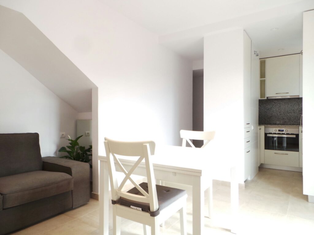 Diseño de interiores moderno en Barcelona: Espacio abierto con sofá gris y mesa blanca Interior de un elegante espacio de planta abierta en Barcelona, con un sofá gris acogedor y una mesa de comedor blanca con sillas a juego, iluminado por luz natural y decorado de manera minimalista.