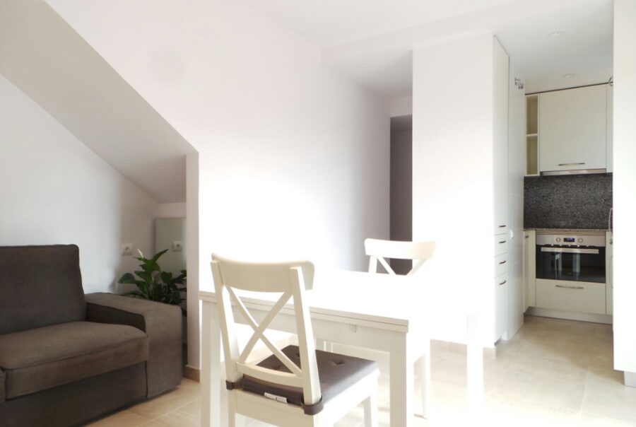 Interior de un elegante espacio de planta abierta en Barcelona, con un sofá gris acogedor y una mesa de comedor blanca con sillas a juego, iluminado por luz natural y decorado de manera minimalista.