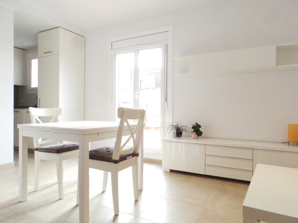 Comedor moderno y minimalista en un proyecto de diseño de interiores en Barcelona, con mesa blanca y sillas, iluminado por grandes ventanas y decorado con un elegante gabinete blanco y plantas.