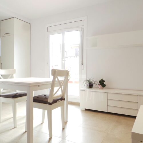 Comedor moderno y minimalista en un proyecto de diseño de interiores en Barcelona, con mesa blanca y sillas, iluminado por grandes ventanas y decorado con un elegante gabinete blanco y plantas.