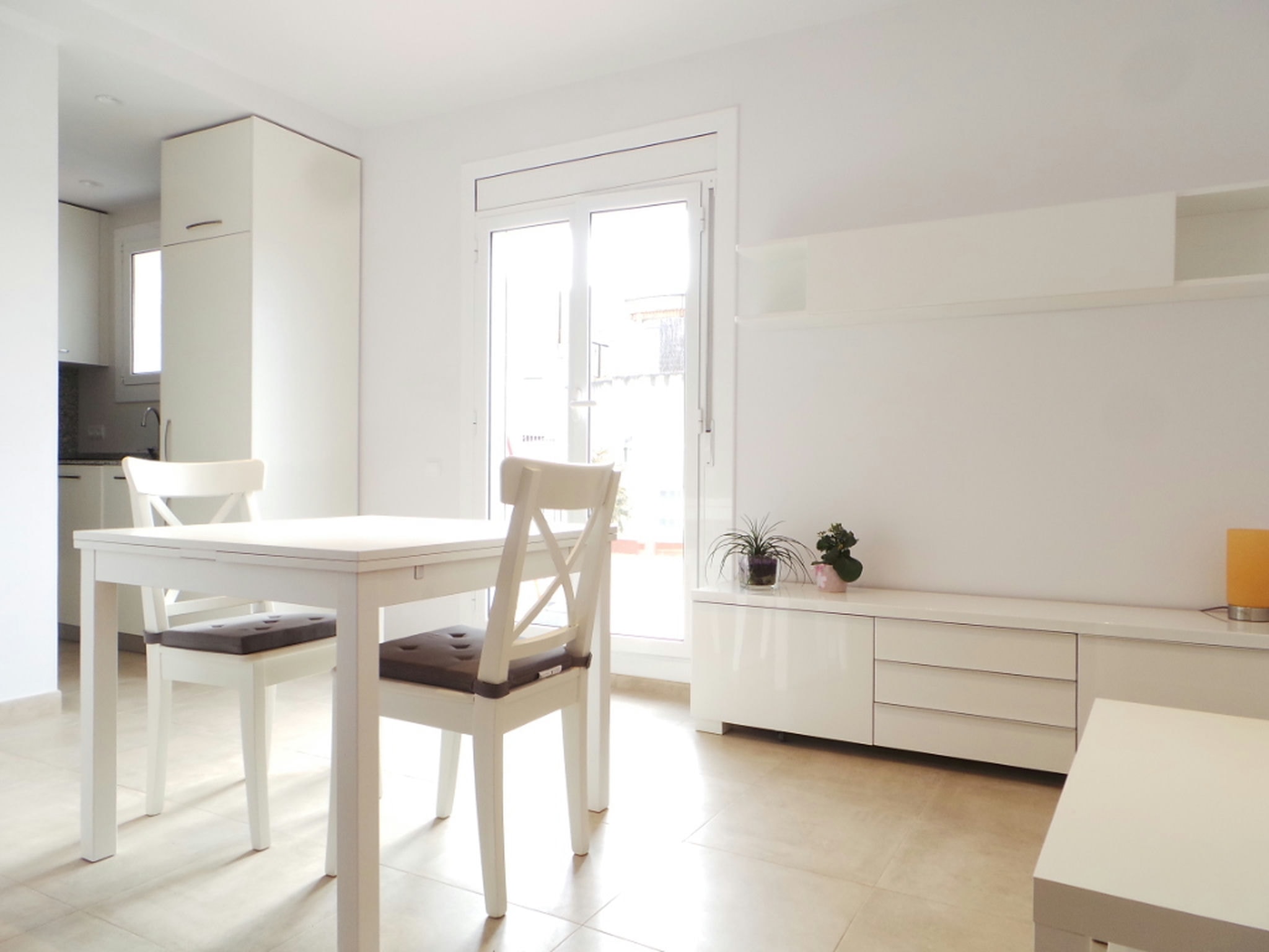 Comedor moderno y minimalista en un proyecto de diseño de interiores en Barcelona, con mesa blanca y sillas, iluminado por grandes ventanas y decorado con un elegante gabinete blanco y plantas.
