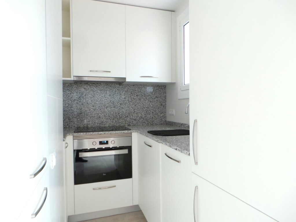 Cocina Moderna con Muebles Blancos y Encimera de Granito en Barcelona Diseño de una cocina moderna en Barcelona con gabinete blanco y encimera de granito pulido, bien iluminada con luz natural.