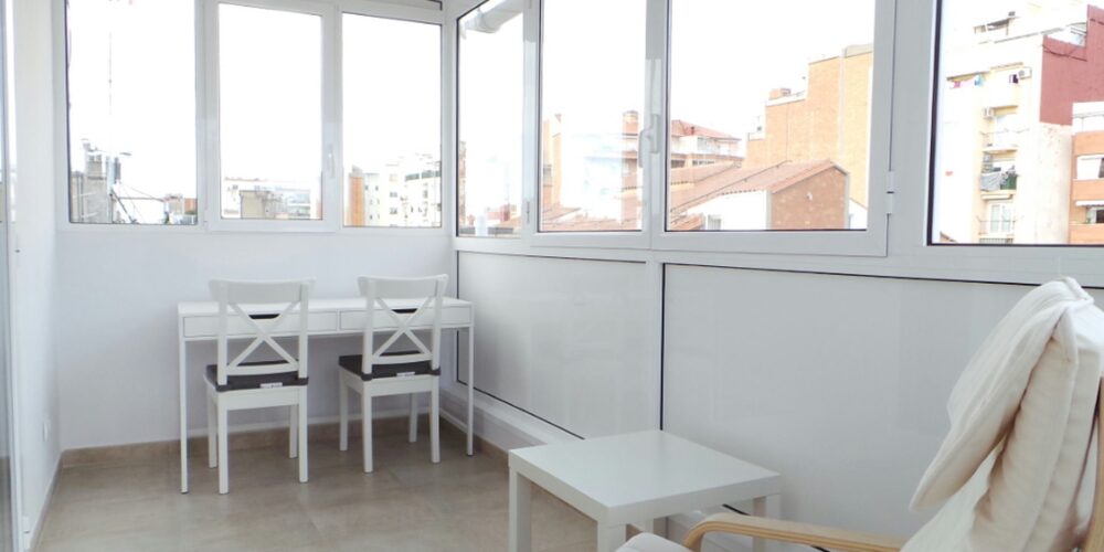 Espacio de diseño de interiores en Barcelona con ventanas de vidrio, área de trabajo con mesas y sillas blancas, sillón cómodo, suelo de baldosa clara y paredes blancas, reflejando la arquitectura contemporánea de alta gama.