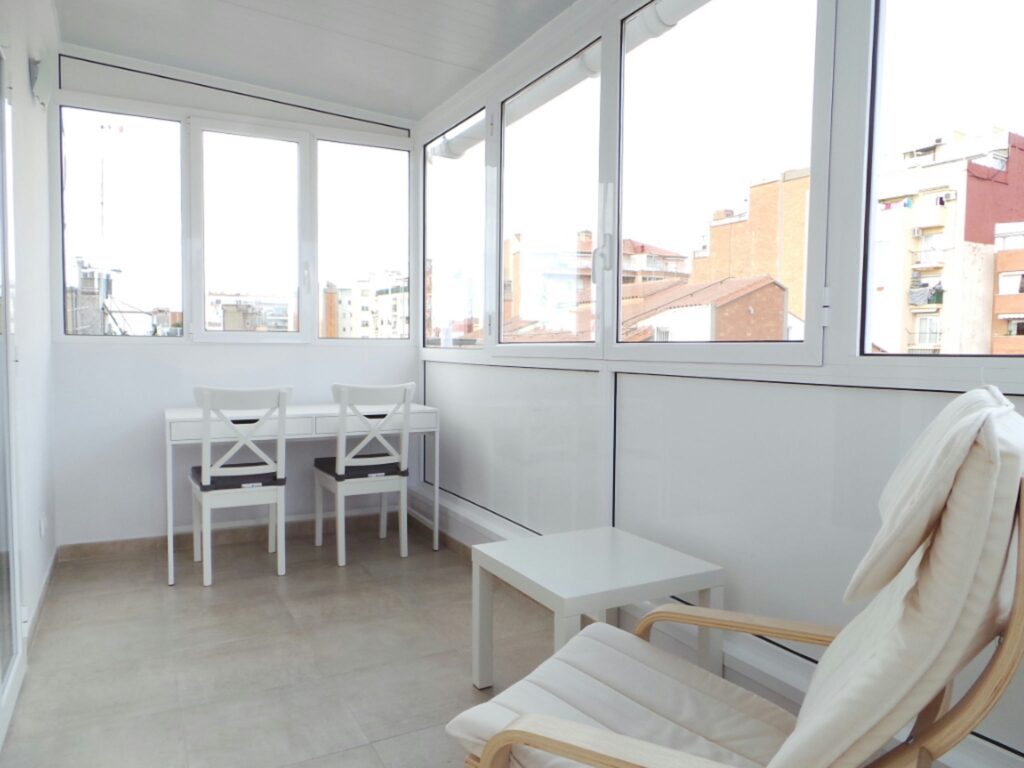 Diseño de interiores moderno en Barcelona con vistas urbanas Espacio de diseño de interiores en Barcelona con ventanas de vidrio, área de trabajo con mesas y sillas blancas, sillón cómodo, suelo de baldosa clara y paredes blancas, reflejando la arquitectura contemporánea de alta gama.