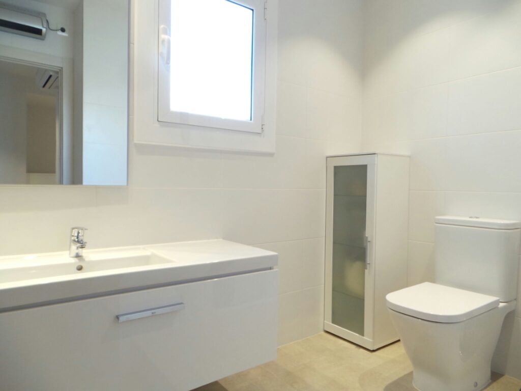 Diseño de baño moderno con materiales de alta calidad en Barcelona Baño contemporáneo en Barcelona con mobiliario minimalista, incluyendo un lavabo elegante, espejo moderno, y un inodoro limpio. La luz natural ilumina el espacio a través de una ventana cercana, mientras un armario de cristal complementa la estética elegante y funcional del diseño.