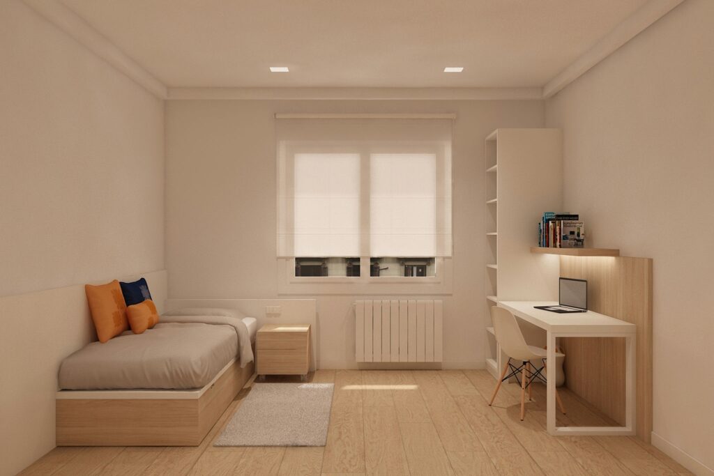 Diseño de interiores moderno en Barcelona: dormitorio acogedor Dormitorio moderno en Barcelona con cama sencilla, cojines naranjas y azules, mesa de noche simple y escritorio con laptop. Espacio minimalista iluminado por luz natural, con suelo de madera y muebles elegantes que reflejan funcionalidad y estilo.