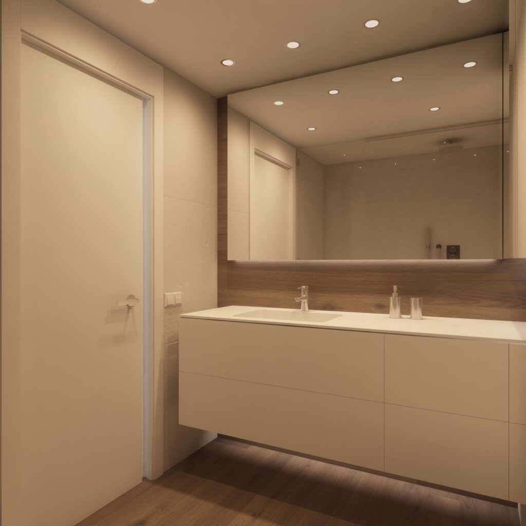 Baño moderno de diseño interior en Barcelona con estética minimalista y funcional Moderno baño minimalista en Barcelona, con mueble de lavabo claro, lavabo de doble seno, grifería elegante, iluminación empotrada y grandes espejos, destacando materiales de calidad y un detalle en madera que aporta calidez.