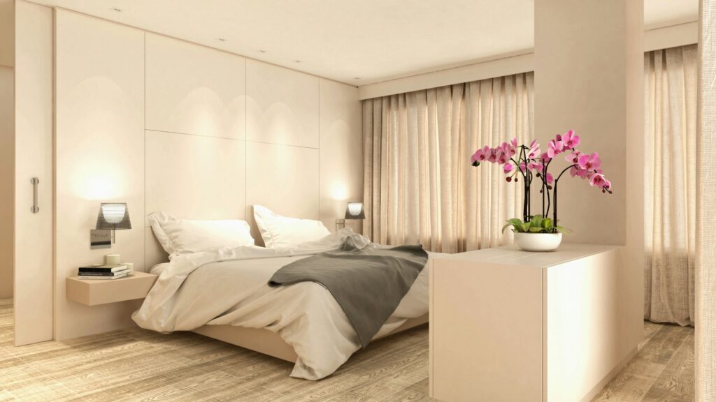 Diseño de interiores de lujo en Barcelona: dormitorio moderno y sofisticado Interior de dormitorio moderno y elegante en Barcelona, con una cama grande, sábanas blancas, mesas de noche minimalistas y una planta orquídea. La luz natural entra a través de cortinas translúcidas, resaltando los materiales de alta calidad como el suelo de madera clara.
