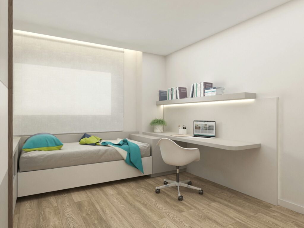 Diseño de interiores moderno en Barcelona: dormitorio elegante y funcional Diseño de interiores de un dormitorio moderno en Barcelona con cama individual, cojines coloridos, escritorio minimalista y una silla ergonómica. Piso de madera clara y luz natural que entra por una gran ventana, resaltando una estética contemporánea.