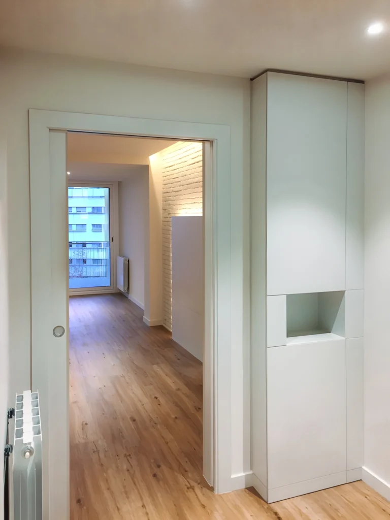 Interior Contemporáneo Minimalista con Luz Natural y Elementos de Diseño Estilizados Un interior contemporáneo minimalista que presenta una puerta que conduce a un área abierta con suelo de madera. La luz natural inunda el espacio a través de una ventana, destacando un alto armario blanco con una sección recortada. Las paredes están pintadas en tonos neutros, con un radiador visible a la izquierda, todo contribuyendo a una atmósfera moderna y aireada.