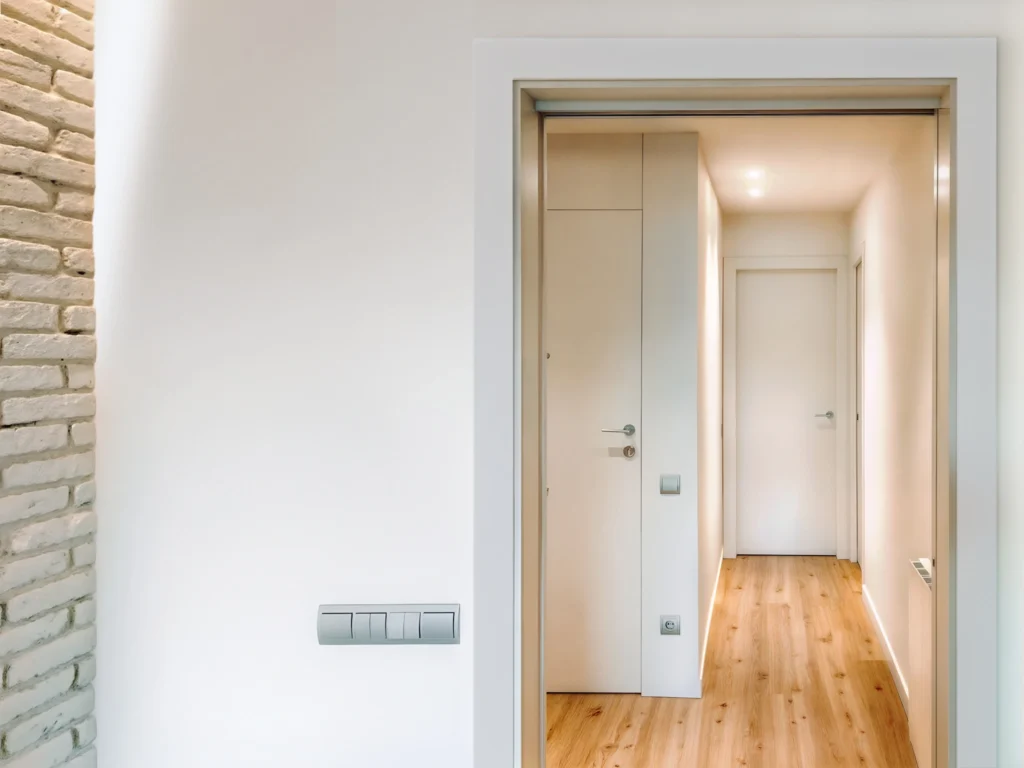 Pasillo Interior Moderno con Puerta Abierta Un pasillo interior limpio y moderno visto a través de una puerta abierta, que presenta una pared blanca texturizada a la izquierda y una puerta de color claro a la derecha. El pasillo brillante tiene una iluminación suave y un suelo de madera clara, con puertas adicionales que alinean el espacio, encarnando una estética de diseño contemporáneo.