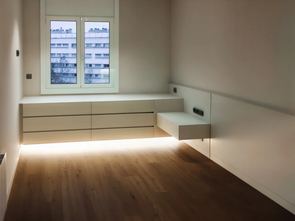 Proyecto de Diseño de Interiores Moderno en Barcelona: Espacio Minimalista de Alta Gama Imagen de un proyecto de diseño de interiores moderno en Barcelona, destacando un espacio minimalista con mobiliario incorporado, iluminación LED sutil y grandes ventanales que permiten la entrada de luz natural sobre suelos de madera clara y paredes de color claro.