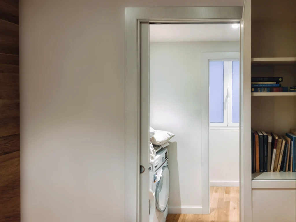 Área de lavandería moderna en proyecto de diseño de interiores de alta gama en Barcelona Vista de un área de lavandería moderna y organizada en un proyecto de diseño interior de lujo en Barcelona, con lavadora, iluminación natural y suelo de madera.