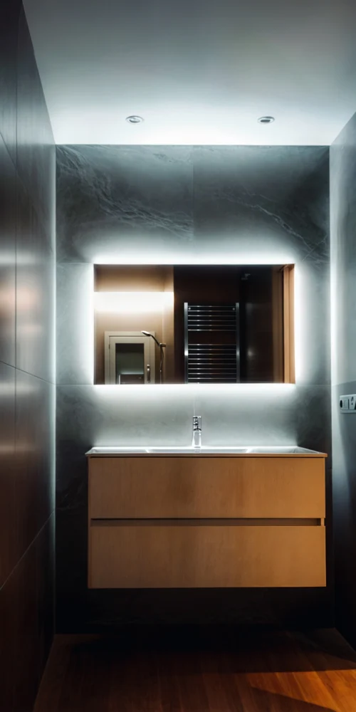 Baño moderno en Barcelona con vanidad de madera clara y paneles oscuros, iluminación suave, espejo iluminado y materiales de mármol, destacando la estética contemporánea y funcionalidad.