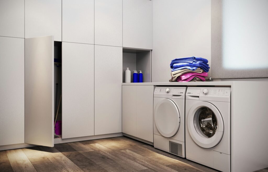 Moderna lavandería de diseño en Barcelona con elementos de alta gama Lavandería contemporánea en Barcelona con mobiliario blanco, dos lavadoras modernas y toallas coloridas sobre una encimera. Suelo de madera y luz natural realzan la estética elegante y funcional del espacio.