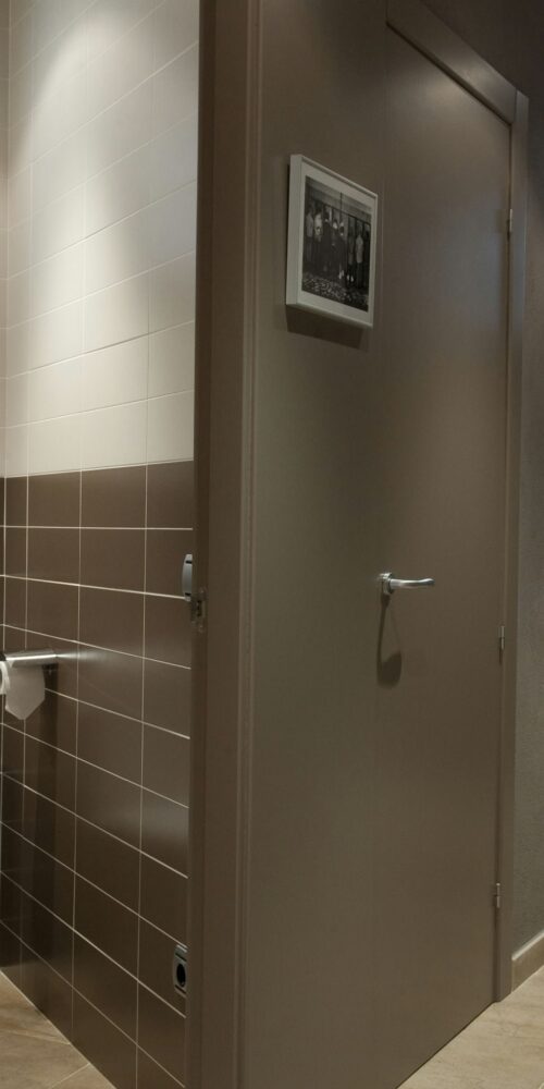 Un baño moderno con una combinación elegante de azulejos claros y oscuros, un inodoro blanco al lado de un dispensador de papel higiénico y luz natural que entra por una ventana.