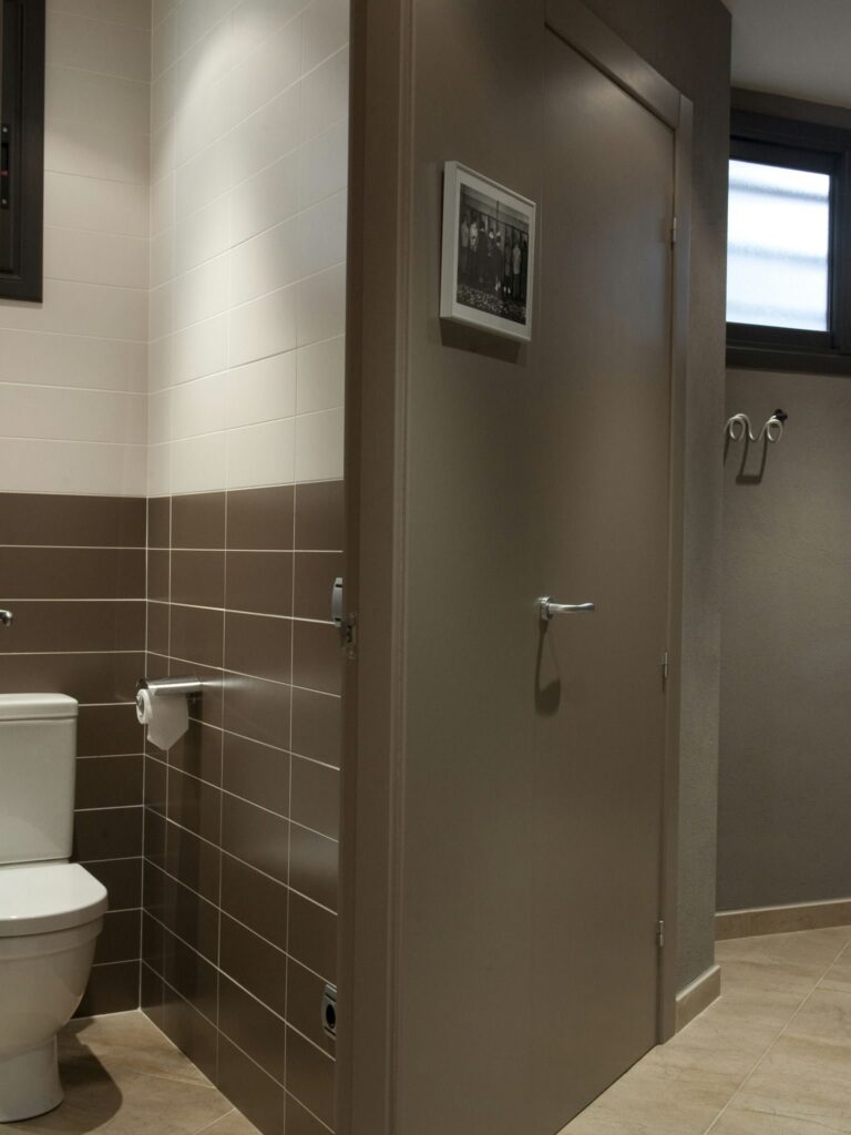 Interior de Baño Contemporáneo con Contraste de Color Estiloso Un baño moderno con una combinación elegante de azulejos claros y oscuros, un inodoro blanco al lado de un dispensador de papel higiénico y luz natural que entra por una ventana.
