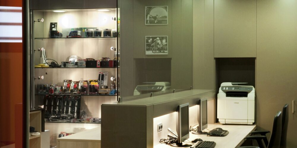 Un espacio de trabajo de oficina moderna, elegante y minimalista, que presenta una estantería iluminada llena de cámaras y accesorios, un escritorio largo con dos configuraciones de computadora y una impresora bien colocada contra la pared.
