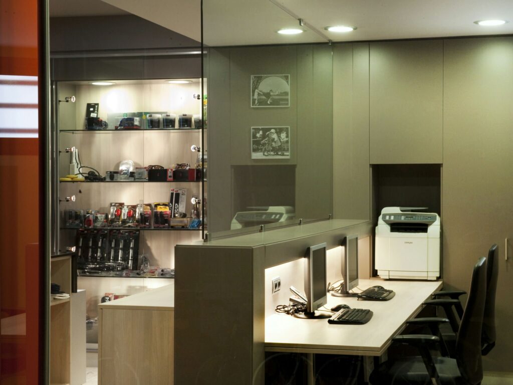Espacio de trabajo de oficina moderna con cámaras y accesorios Un espacio de trabajo de oficina moderna, elegante y minimalista, que presenta una estantería iluminada llena de cámaras y accesorios, un escritorio largo con dos configuraciones de computadora y una impresora bien colocada contra la pared.