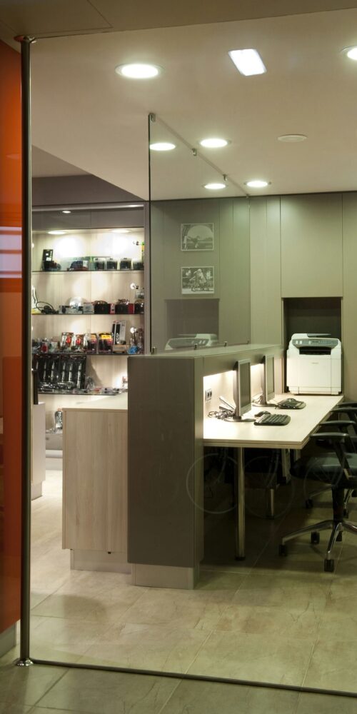 Un interior de oficina contemporáneo con un tabique de cristal, estaciones de trabajo elegantes con computadoras, sillas ergonómicas y una impresora. Estantes llenos de cámaras y accesorios de fotografía crean una atmósfera profesional.