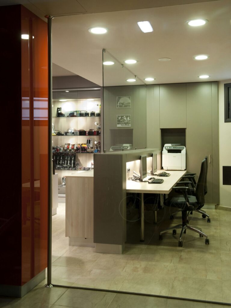 Espacio de Oficina Moderno con Tabique de Cristal y Estaciones de Trabajo Ergonómicas Un interior de oficina contemporáneo con un tabique de cristal, estaciones de trabajo elegantes con computadoras, sillas ergonómicas y una impresora. Estantes llenos de cámaras y accesorios de fotografía crean una atmósfera profesional.