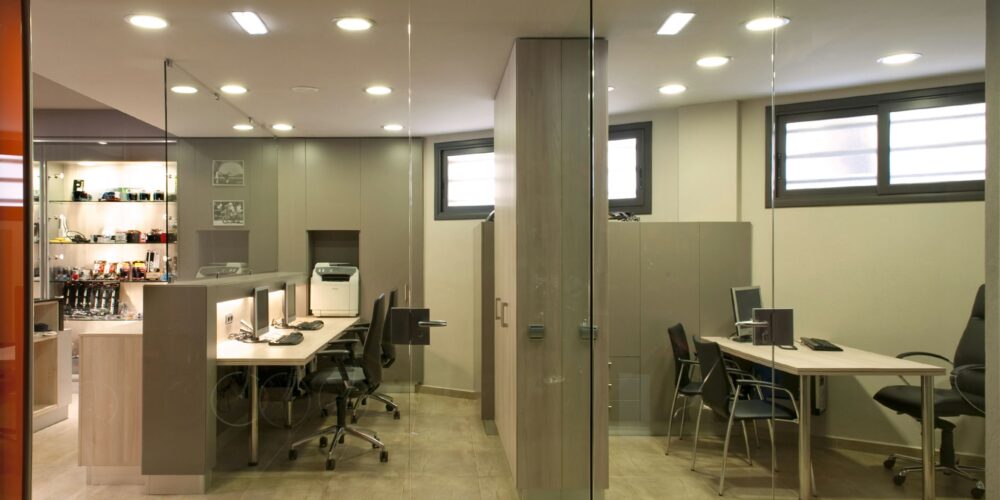 Un entorno de oficina contemporáneo que presenta paredes de vidrio, escritorios de madera, sillas ergonómicas y una iluminación natural brillante, mostrando un espacio de trabajo organizado y funcional.