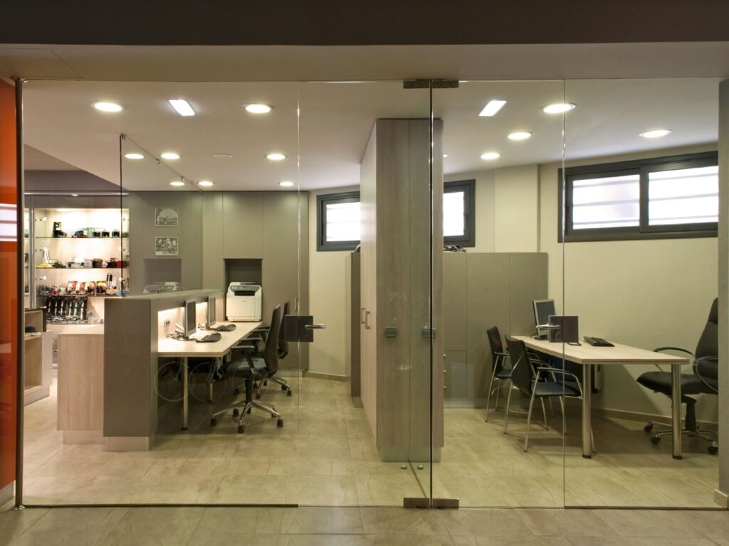 Diseño de Espacio de Oficina Moderna con Paredes de Vidrio Abierto y Estaciones de Trabajo Ergonómicas Un entorno de oficina contemporáneo que presenta paredes de vidrio, escritorios de madera, sillas ergonómicas y una iluminación natural brillante, mostrando un espacio de trabajo organizado y funcional.