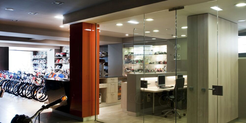 Interior de una oficina contemporánea con una sección de paredes de vidrio, exhibiendo prominentemente bicicletas a la izquierda y una disposición de oficina organizada a la derecha con escritorios, sillas y estanterías elegantes, todo destacado por una iluminación abundante y un diseño elegante.
