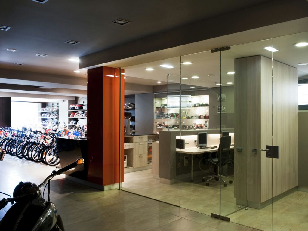 Espacio de oficina moderno con paredes de vidrio y exhibición de bicicletas Interior de una oficina contemporánea con una sección de paredes de vidrio, exhibiendo prominentemente bicicletas a la izquierda y una disposición de oficina organizada a la derecha con escritorios, sillas y estanterías elegantes, todo destacado por una iluminación abundante y un diseño elegante.
