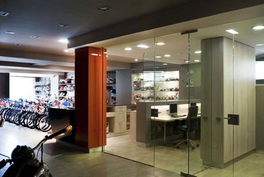 Interior de una oficina contemporánea con una sección de paredes de vidrio, exhibiendo prominentemente bicicletas a la izquierda y una disposición de oficina organizada a la derecha con escritorios, sillas y estanterías elegantes, todo destacado por una iluminación abundante y un diseño elegante.
