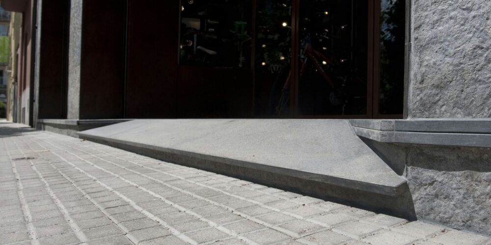 Entrada de edificio moderno con una rampa de concreto y pavimento de piedra con patrones bajo una iluminación brillante.