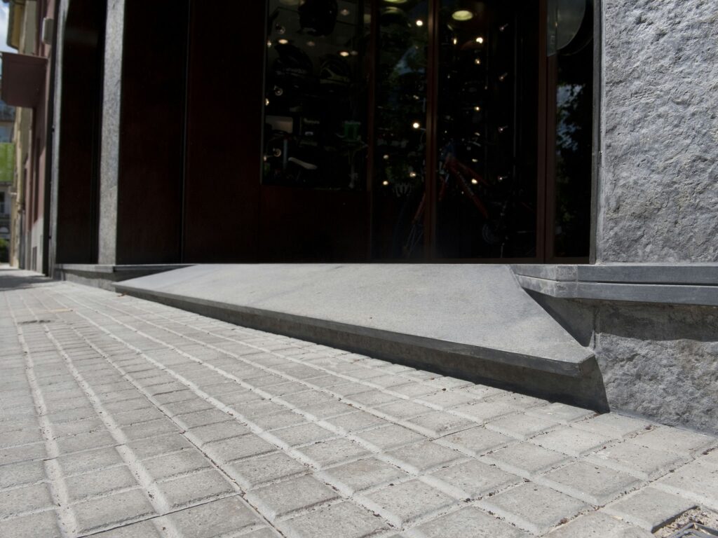 Entrada de Edificio Contemporáneo con Rampa de Concreto Accesible Entrada de edificio moderno con una rampa de concreto y pavimento de piedra con patrones bajo una iluminación brillante.
