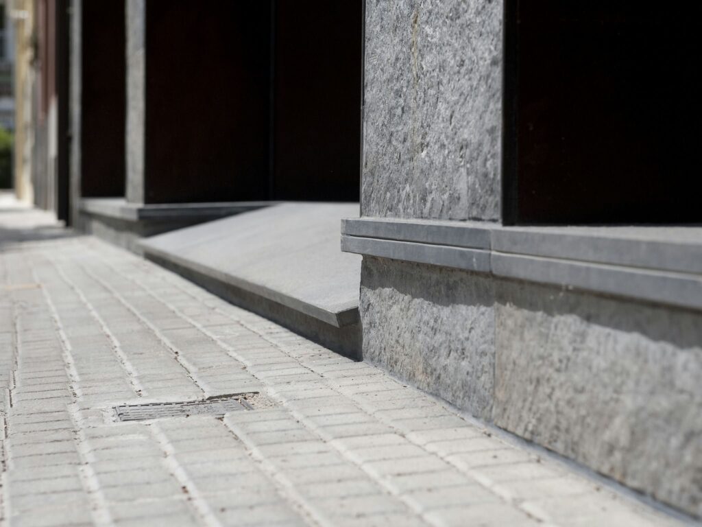 Primer Plano de una Superficie Pavimentada en la Transición entre la Acera y la Entrada del Edificio Vista detallada de un camino de piedra texturizada que presenta losas de pavimento rectangulares en el área de transición entre una acera y la entrada de un edificio moderno, con una pared de piedra gris en el fondo y luz natural creando sombras sutiles.