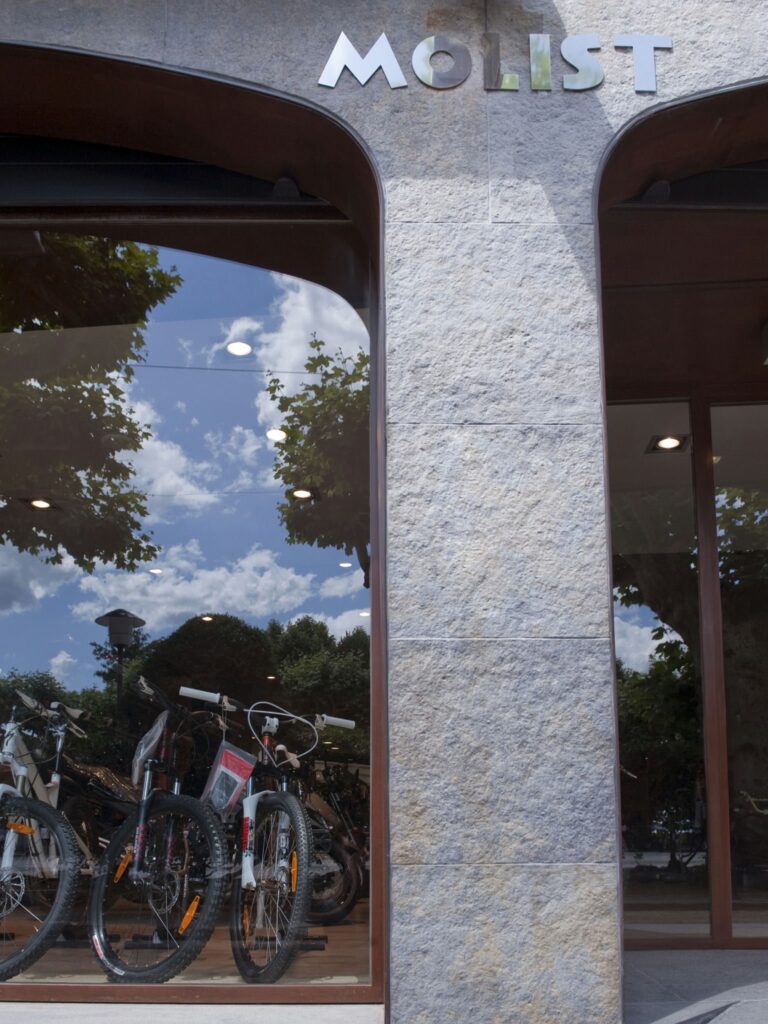 MOLIST Tienda de Bicicletas: Destino de Ciclismo Urbano Brillante escaparate de MOLIST que presenta varias bicicletas en exhibición a través de grandes ventanales de cristal, reflejando árboles y nubes. La arquitectura moderna de la tienda incluye bordes redondeados y un exterior similar a la piedra, enfatizando un vibrante estilo de vida ciclista urbano.