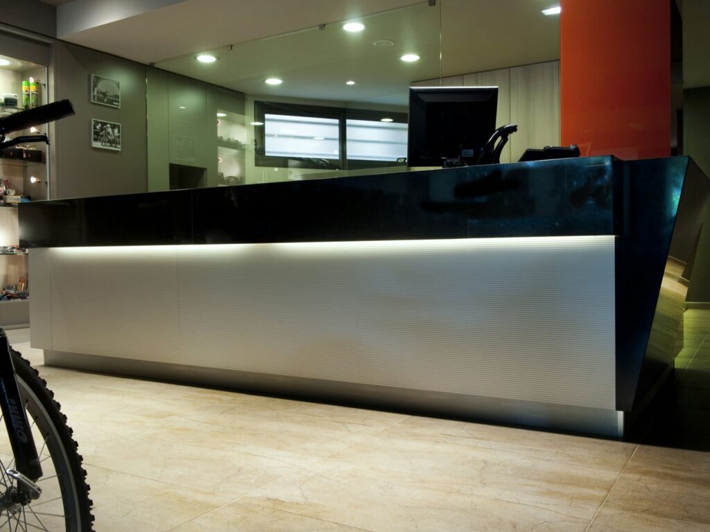 Diseño de Recepción Moderna en Barcelona: Funcionalidad y Estética Moderna recepción en un proyecto de diseño de interiores de alta gama en Barcelona, con un escritorio elegante que combina materiales oscuros y acabados iluminados, destacando la funcionalidad y estética. El espacio presenta suelos de mármol y texturas detalladas, reflejando estilos arquitectónicos contemporáneos. Una bicicleta se vislumbra en primer plano, añadiendo un toque único al entorno.