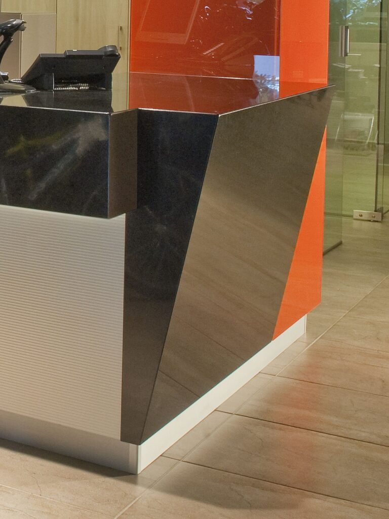 escritorio de recepción moderno y elegante con diseño geométrico Un escritorio de recepción moderno con un diseño geométrico que presenta una superficie brillante negra prominente y una sección blanca texturizada acentuada por naranja. El interior contemporáneo exhibe paneles de madera y una puerta de vidrio.