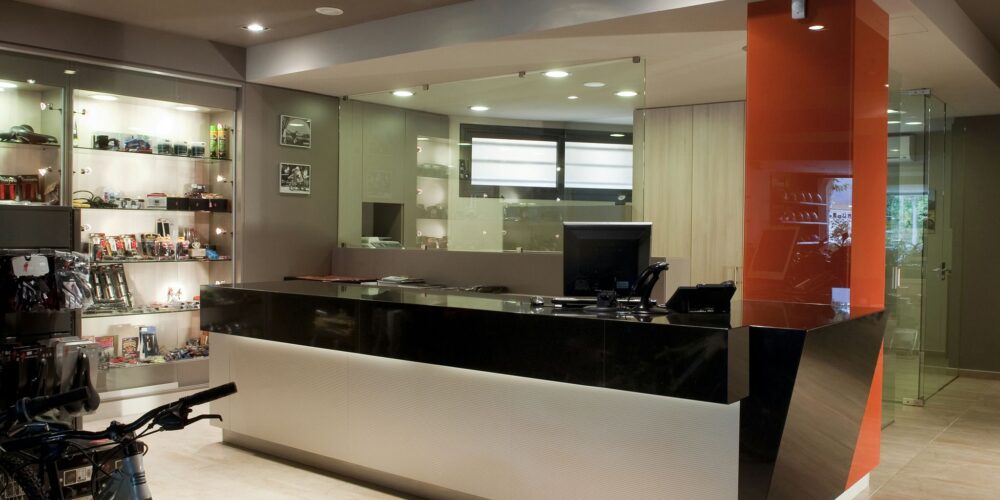 Una área de recepción contemporánea que muestra un mostrador estiloso en blanco y negro con un llamativo acento naranja. El mostrador está equipado con una computadora y un teléfono, y presenta estanterías de vidrio llenas de varios gadgets y accesorios. El espacio bien iluminado, destacado por iluminación empotrada y un suelo pulido, también incluye dos bicicletas aparcadas a la izquierda, enfatizando una atmósfera dinámica y moderna.