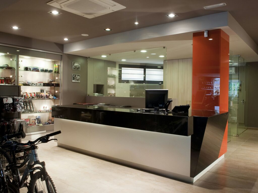 Área de Recepción Moderna con Diseño Elegante y Acabados de Estilo de Vida Activo Una área de recepción contemporánea que muestra un mostrador estiloso en blanco y negro con un llamativo acento naranja. El mostrador está equipado con una computadora y un teléfono, y presenta estanterías de vidrio llenas de varios gadgets y accesorios. El espacio bien iluminado, destacado por iluminación empotrada y un suelo pulido, también incluye dos bicicletas aparcadas a la izquierda, enfatizando una atmósfera dinámica y moderna.