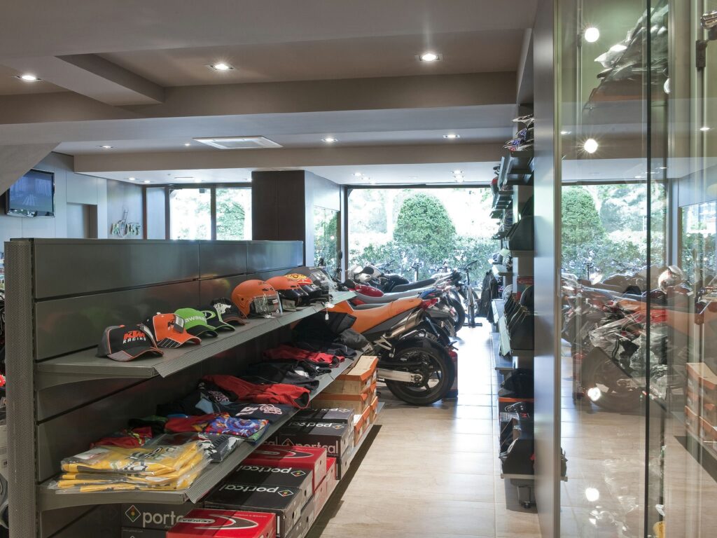 Tienda Moderna de Accesorios para Motocicletas con Equipos y Motocicletas de Marca Interior de una tienda de accesorios para motocicletas que cuenta con estantes organizados con gorras y ropa, iluminación brillante y motocicletas visibles a través de grandes ventanas con vegetación afuera, mostrando un espacio comercial acogedor para entusiastas de las motocicletas.